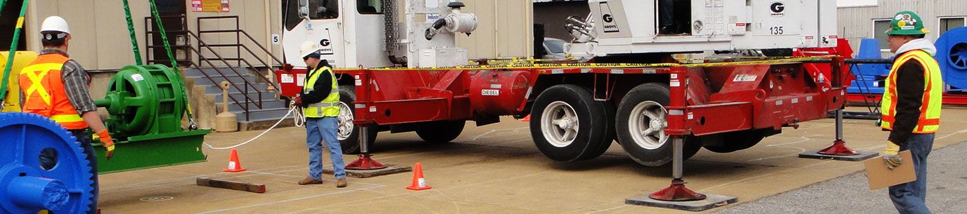 An outdoor practical examination at an ITI training center where a trainee is guiding a large green winch onto a transport trailer, emphasizing real-world rigging and transport safety scenarios.